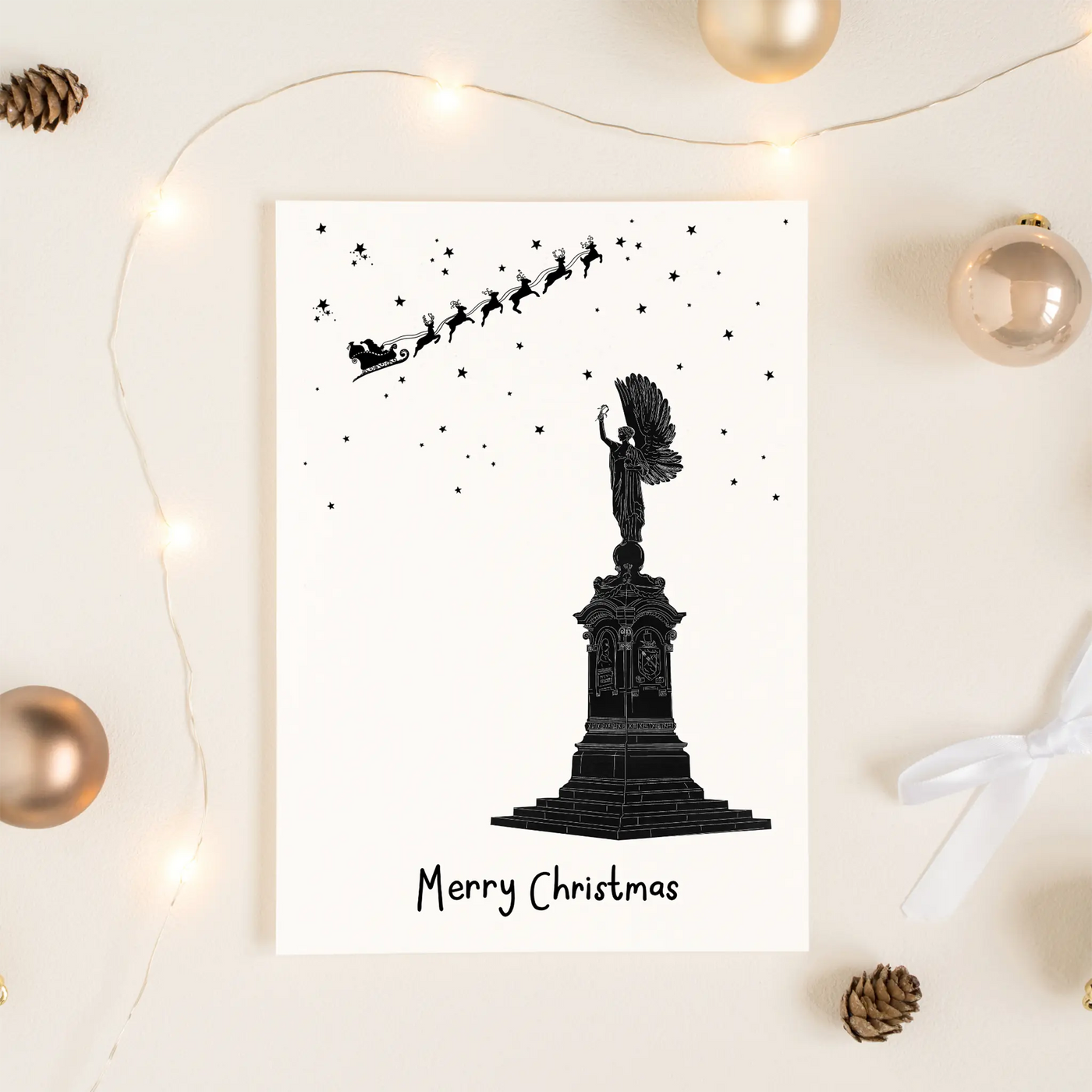 A Christmas greetings cards onto of a brown Kraft envelope. The card depicts a hand illustrated drawing of the Brighton Peace Statue. An angel stand atop a plinth with its arm raise. Overhead smalls stars and Santas sleigh and reindeer can be seen up high.