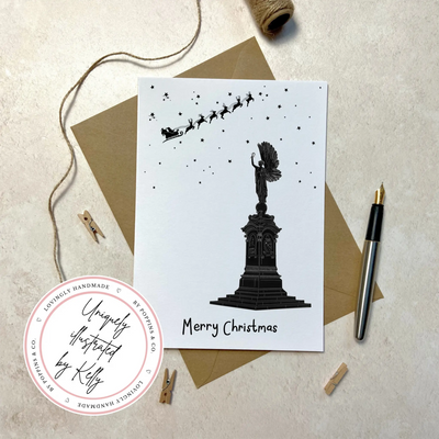 A Christmas greetings cards onto of a brown Kraft envelope. The card depicts a hand illustrated drawing of the Brighton Peace Statue. An angel stand atop a plinth with its arm raise. Overhead smalls stars and Santas sleigh and reindeer can be seen up high.