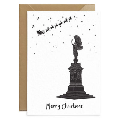 A Christmas greetings cards on top of a brown Kraft envelope in front of a white background. The card depicts a hand illustrated drawing of the Brighton Peace Statue. An angel stand atop a plinth with its arm raise. Overhead smalls stars and Santas sleigh and reindeer can be seen up high.