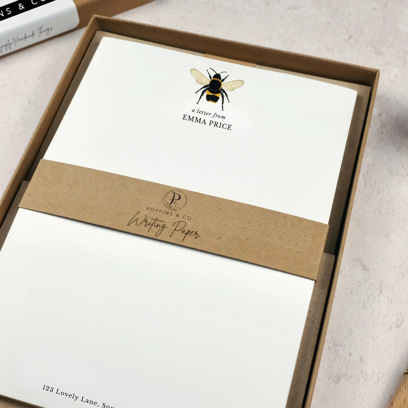 A personalised bumblebee letter writing set by Poppins and co in a brown kraft luxury gift box with a cardboard band around the paper within the box. The writing paper is white and has an illustration of a bee at the top of the page. Italic script reads 'a letter from' with the name 'Emma Price' typed in all caps font below it.