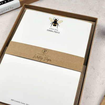 A personalised bumblebee letter writing set by Poppins and co in a brown kraft luxury gift box with a cardboard band around the paper within the box. The writing paper is white and has an illustration of a bee at the top of the page. Italic script reads 'a letter from' with the name 'Emma Price' typed in all caps font below it.