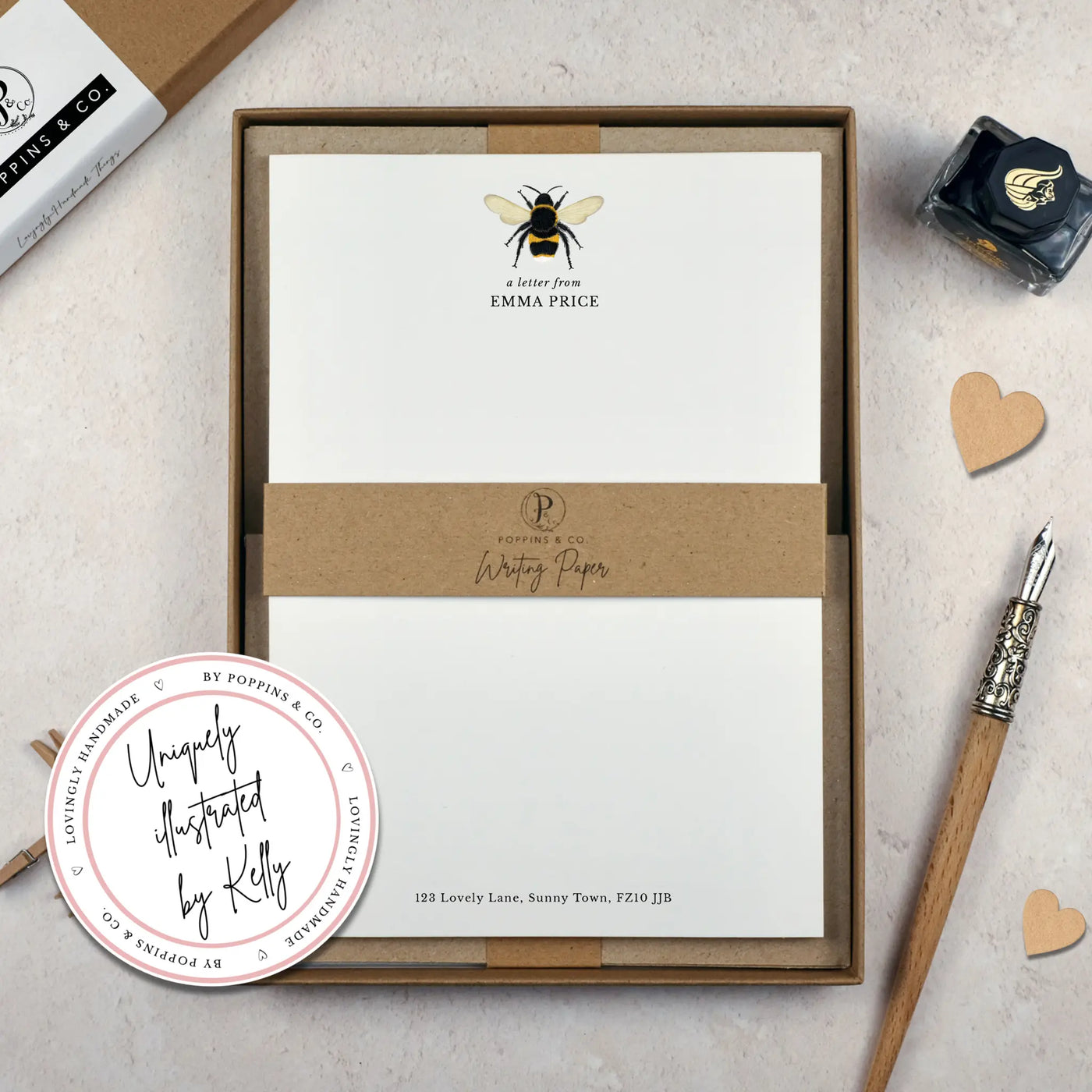 A personalised bumblebee letter writing set by Poppins and co in a brown kraft luxury gift box with a cardboard band around the paper within the box. The writing paper is white and has an illustration of a bee at the top of the page. Italic script reads 'a letter from' with the name 'Emma Price' typed in all caps font below it. At the bottom of the page is a typed address.
