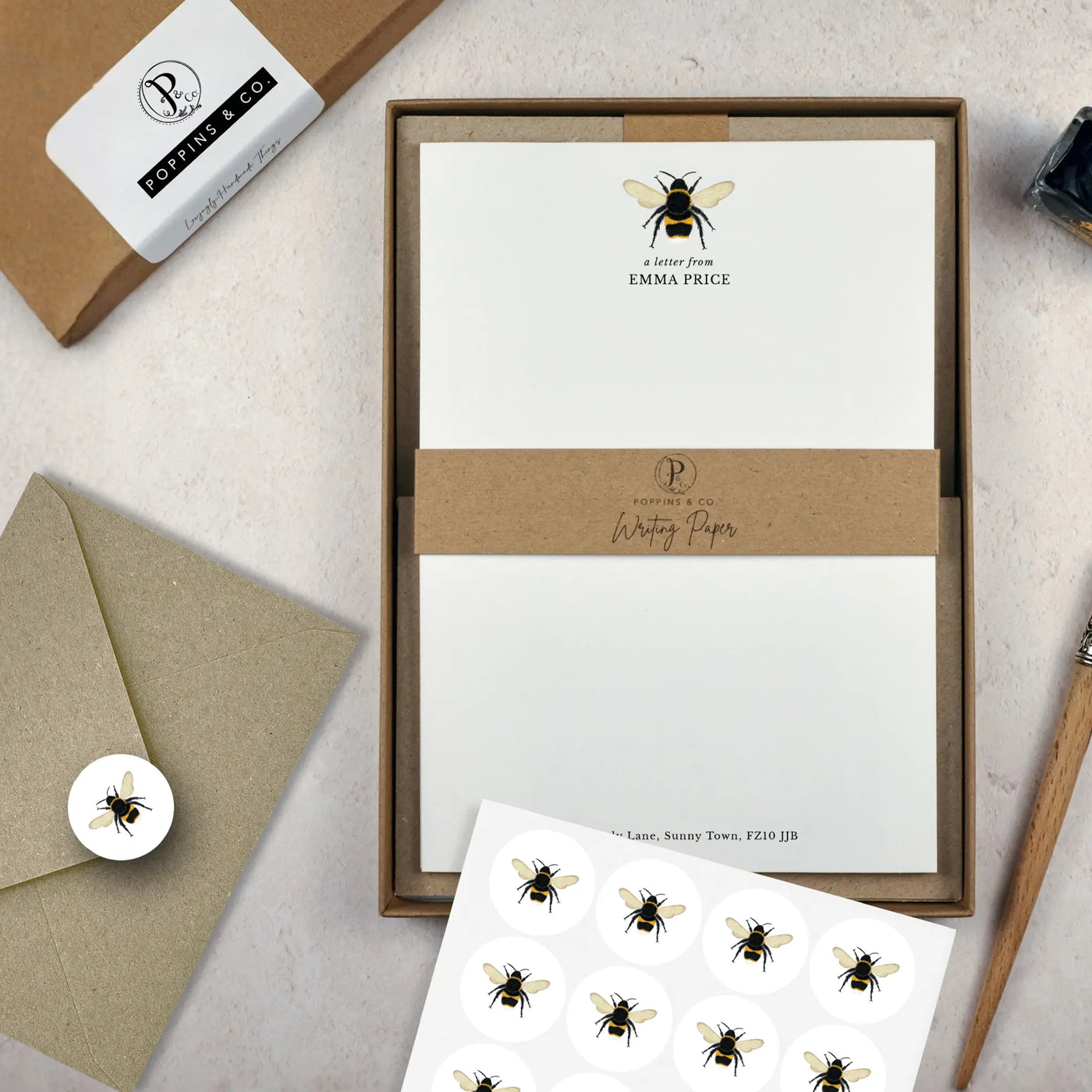 A bumblebee letter writing set by in a brown kraft luxury gift box with a cardboard band around the paper within the box. The writing paper is white and has an illustration of a bumblebee at the top of the page. Italic script reads 'a letter from' with the name 'Emma Price' typed in all caps font below it. On top of the box lays a sheet of round envelope stickers with the same bee illustration on each. To the left lays a brown kraft envelope with an envelope sticker seal on it.