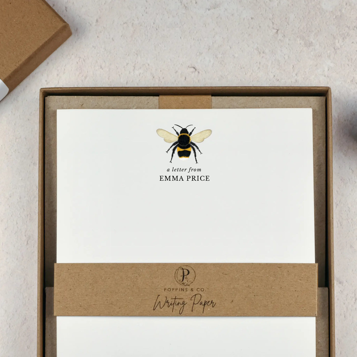 A personalised bumblebee letter writing set by Poppins and co in a brown kraft luxury gift box with a cardboard band around the paper within the box. The writing paper is white and has an illustration of a bee at the top of the page. Italic script reads 'a letter from' with the name 'Emma Price' typed in all caps font below it.