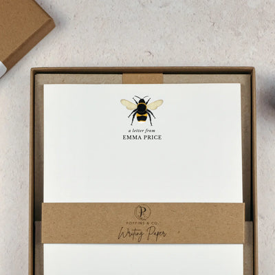 A personalised bumblebee letter writing set by Poppins and co in a brown kraft luxury gift box with a cardboard band around the paper within the box. The writing paper is white and has an illustration of a bee at the top of the page. Italic script reads 'a letter from' with the name 'Emma Price' typed in all caps font below it.