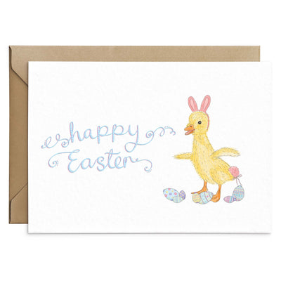 A funny Easter Greetings Card featuring a duckling wearing pink easter bunny ears and a pink bunny tail. Pastel coloured pattern easter eggs are scattered below the ducklings feet. Pink and blue scripted writing above reads 'Happy Easter'. White background greetings card with a brown Kraft envelope laying behind. Greetings card by Poppins and Co.