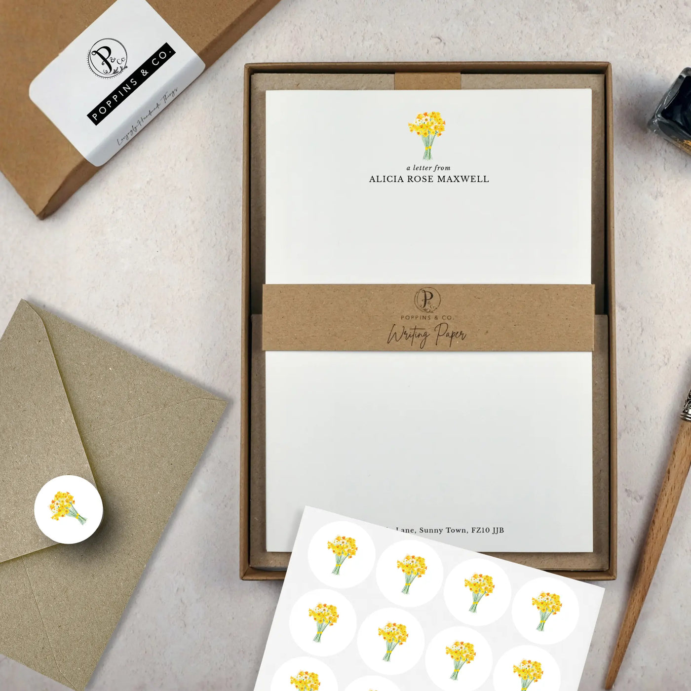 A letter writing set by in a brown kraft luxury gift box with a cardboard band around the paper within the box. The writing paper is white and has a small bunch of yellow daffodils illustrated at the top of the page. Italic script reads 'a letter from' with the name 'Alicia Rose Maxwell' typed in all caps font below it. On top of the box lays a sheet of round envelope stickers with the same daffodil illustration on each. To the left lays a brown kraft envelope with an envelope sticker seal on it.