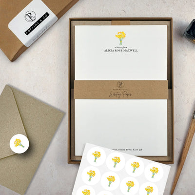 A letter writing set by in a brown kraft luxury gift box with a cardboard band around the paper within the box. The writing paper is white and has a small bunch of yellow daffodils illustrated at the top of the page. Italic script reads 'a letter from' with the name 'Alicia Rose Maxwell' typed in all caps font below it. On top of the box lays a sheet of round envelope stickers with the same daffodil illustration on each. To the left lays a brown kraft envelope with an envelope sticker seal on it.