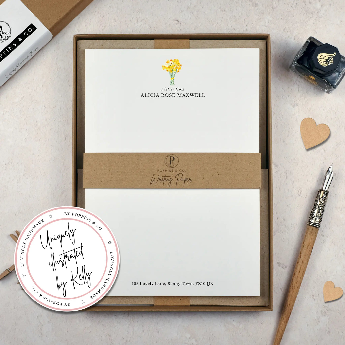 A personalised letter writing set by Poppins and co in a brown kraft luxury gift box with a cardboard band around the paper within the box. The writing paper is white and has a small bunch of yellow daffodils illustrated at the top of the page. Italic script reads 'a letter from' with the name 'Alicia Rose Maxwell' typed in all caps font below it. At the bottom of the page is a typed address.