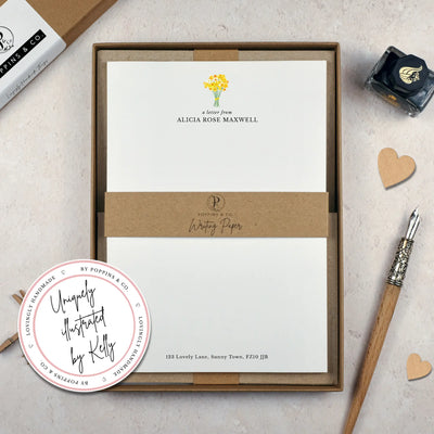 A personalised letter writing set by Poppins and co in a brown kraft luxury gift box with a cardboard band around the paper within the box. The writing paper is white and has a small bunch of yellow daffodils illustrated at the top of the page. Italic script reads 'a letter from' with the name 'Alicia Rose Maxwell' typed in all caps font below it. At the bottom of the page is a typed address.