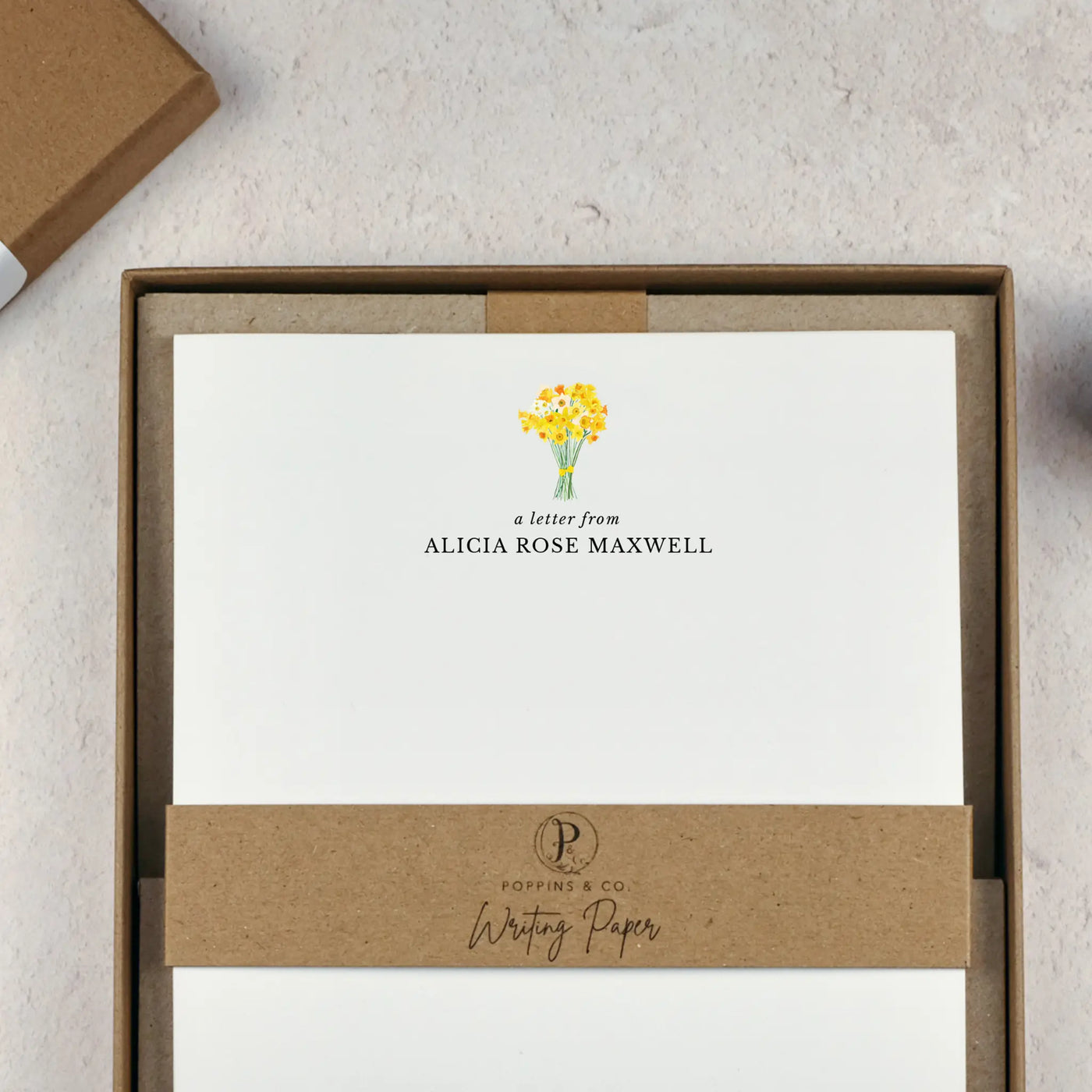 A personalised letter writing set by Poppins and co in a brown kraft luxury gift box with a cardboard band around the paper within the box. The cardboard band reads Poppins & Co - Writing Paper. The writing paper is white and has a small bunch of yellow daffodils illustrated at the top of the page. Italic script reads 'a letter from' with the name 'Alicia Rose Maxwell' typed in all caps font below it. 