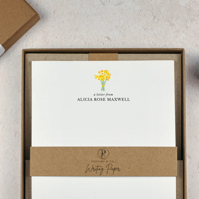 A personalised letter writing set by Poppins and co in a brown kraft luxury gift box with a cardboard band around the paper within the box. The cardboard band reads Poppins & Co - Writing Paper. The writing paper is white and has a small bunch of yellow daffodils illustrated at the top of the page. Italic script reads 'a letter from' with the name 'Alicia Rose Maxwell' typed in all caps font below it. 