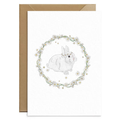 A spring themed greetings cards featuring a white bunny wearing a daisy crown pictured inside a rather made of daisies and greenery. No text. Photo shows one cards laid out on top of recycled brown envelope. Easter greetings card by Poppins and co.