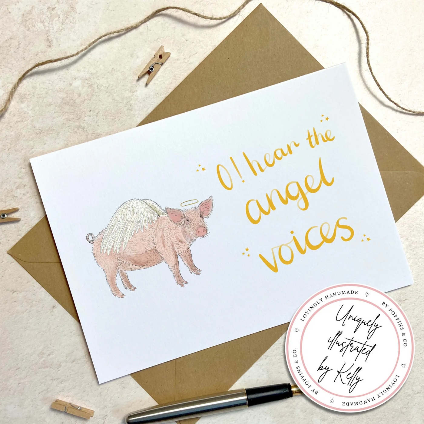 A unique christmas card lays on a table on top of a brown envelope. The card depicts a whimsical illustration of cute pig with angel wings and a halo.  Hand scripted yellow text reads 'O! hear the angel voices'.