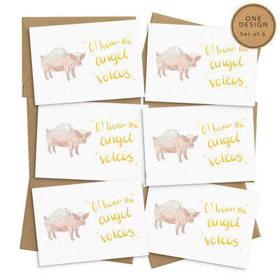 A unique set of 6 christmas cards lays on white background with 6 brown envelopes. The card depicts a whimsical illustration of cute pig with angel wings and a halo.  Hand scripted yellow text reads 'O! hear the angel voices'.