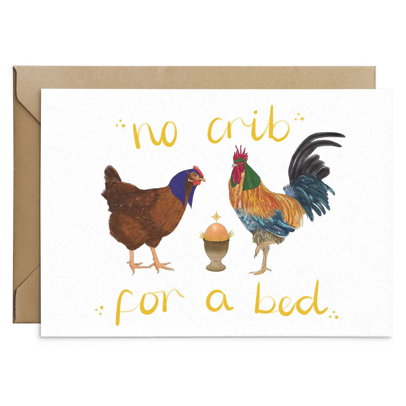 A unique christmas cards lays on a white background with a brown envelope. The card design depicts a whimsical illustration of nativity scene with Chicken as Mary, a cockerel as Joseph and an egg in an egg . Hand scripted yellow text reads 'no crib for a bed'.