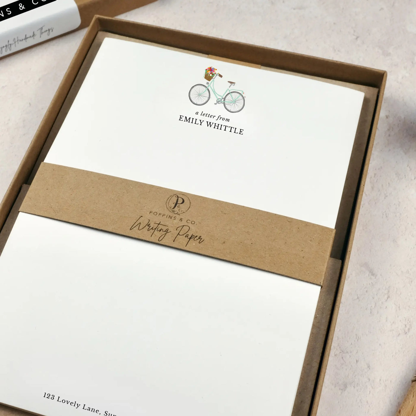 A personalised bicycle letter writing set by Poppins and co in a brown kraft luxury gift box with a cardboard band around the paper within the box. The writing paper is white and has an illustration of a bicycle with flowers in the basket at the top of the page. Italic script reads 'a letter from' with the name 'Emily Whittle' typed in all caps font below it. At the bottom of the page is a typed address.