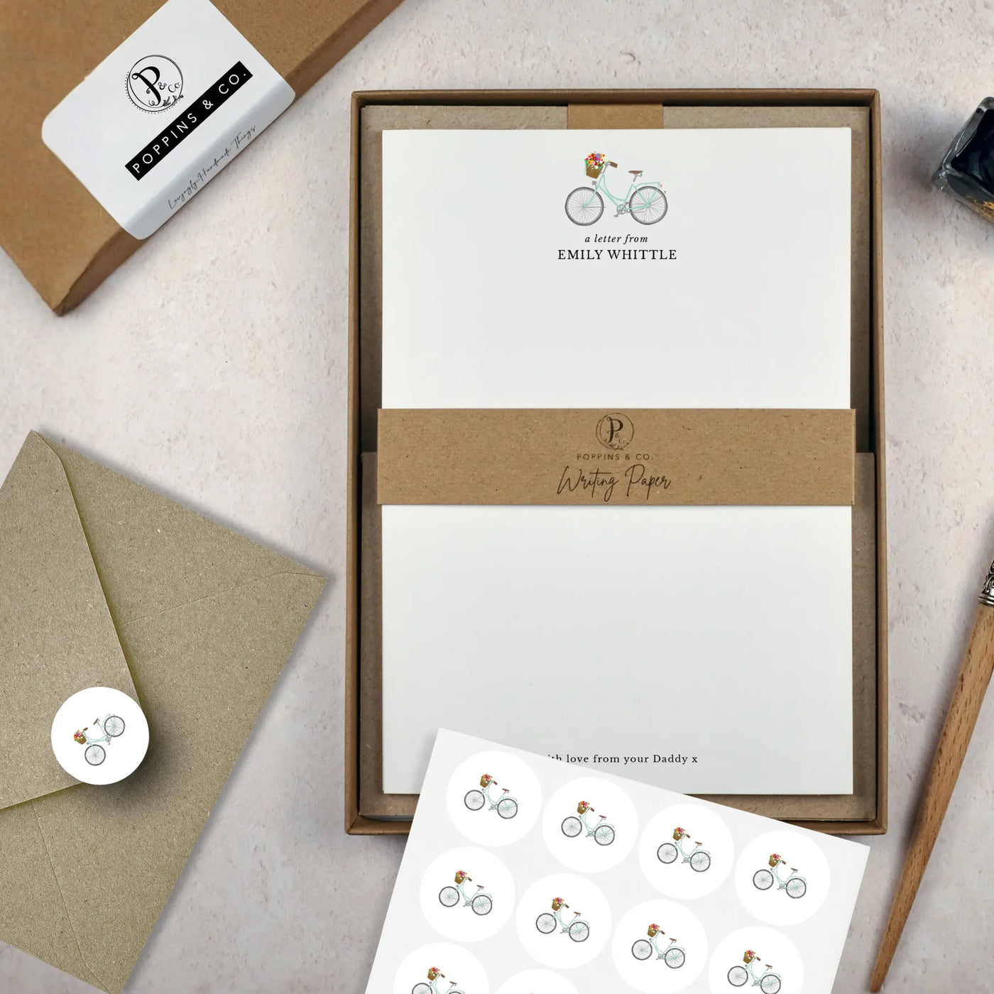 A floral bicycle writing set by in a brown kraft luxury gift box with a cardboard band around the paper within the box. The writing paper is white and has an illustration of a pale blue green bicycle with flowers at the top of the page. Italic script reads 'a letter from' with the name 'Emily Whittle' typed in all caps font below it. On top of the box lays a sheet of round envelope stickers with the same bike illustration on each. To the left lays a brown kraft envelope with an envelope sticker seal on it.