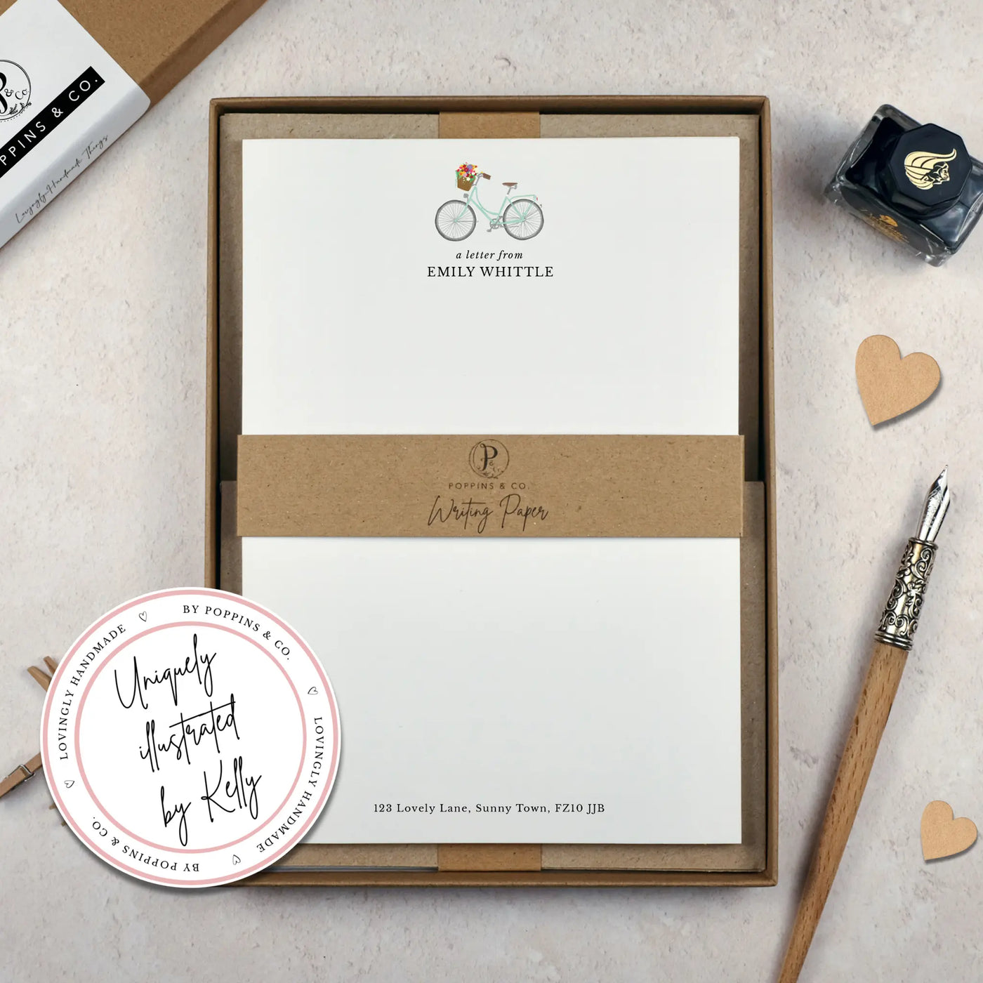 A personalised bicycle letter writing set by Poppins and co in a brown kraft luxury gift box with a cardboard band around the paper within the box. The writing paper is white and has an illustration of a bicycle with flowers in the basket at the top of the page. Italic script reads 'a letter from' with the name 'Emily Whittle' typed in all caps font below it. At the bottom of the page is a typed address.