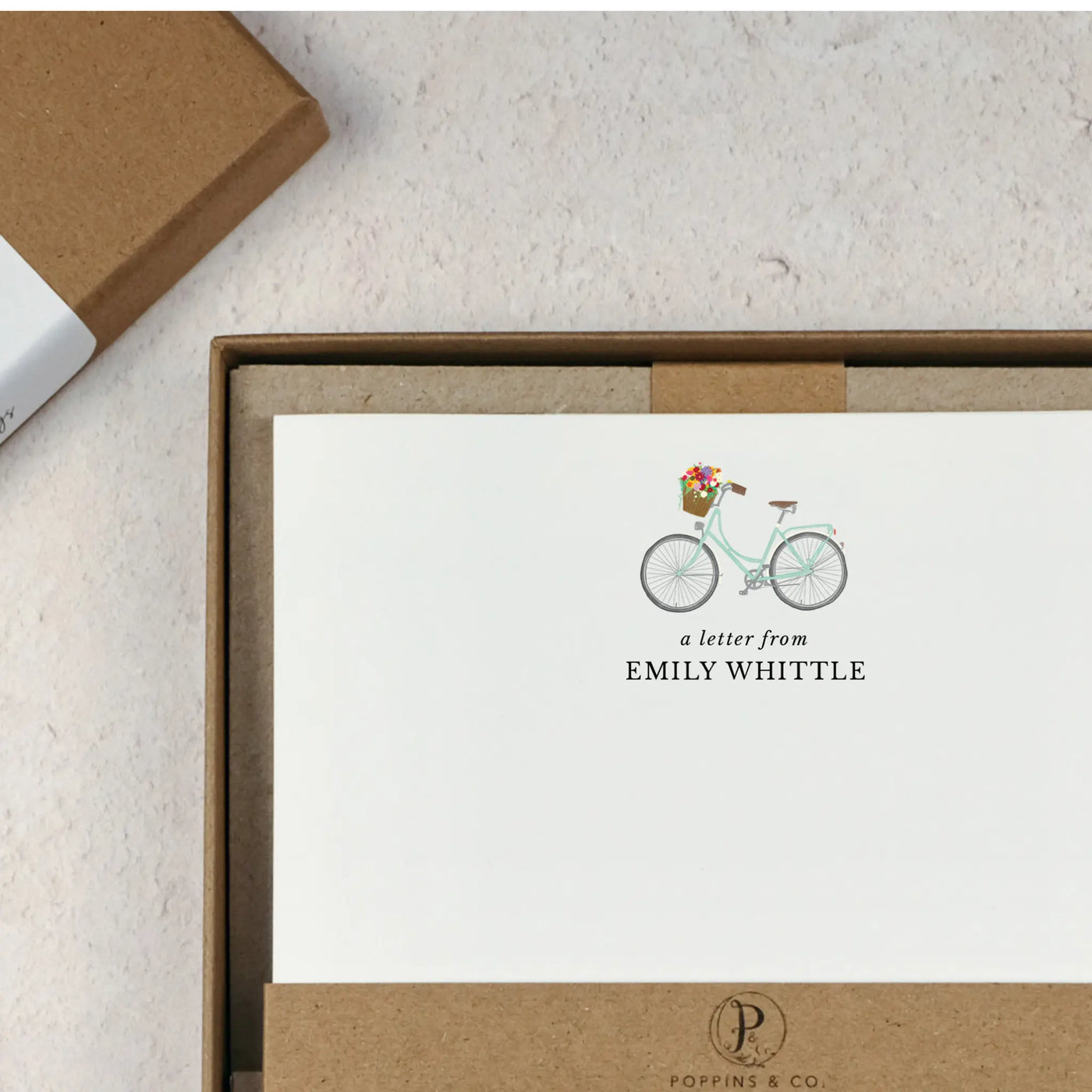 A personalised bicycle letter writing set by Poppins and co in a brown kraft luxury gift box with a cardboard band around the paper within the box. The writing paper is white and has an illustration of a bicycle with flowers in the basket at the top of the page. Italic script reads 'a letter from' with the name 'Emily Whittle' typed in all caps font below it. At the bottom of the page is a typed address.