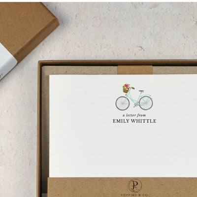 A personalised bicycle letter writing set by Poppins and co in a brown kraft luxury gift box with a cardboard band around the paper within the box. The writing paper is white and has an illustration of a bicycle with flowers in the basket at the top of the page. Italic script reads 'a letter from' with the name 'Emily Whittle' typed in all caps font below it. At the bottom of the page is a typed address.