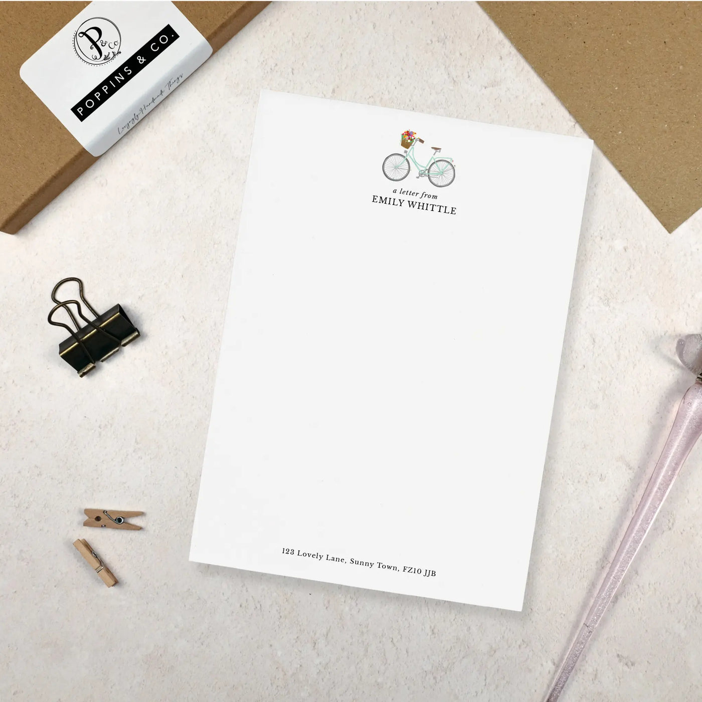 A single sheet of personalised bicycle writing paper. The writing paper is white and has an illustration of a bicycle with flowers in the basket at the top of the page. Italic script reads 'a letter from' with the name 'Emily Whittle' typed in all caps font below it. At the bottom of the page is a typed address.