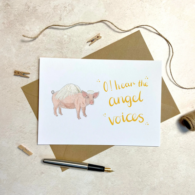 A unique christmas card lays on a table. The card depicts a whimsical illustration of cute pig with angel wings and a halo.  Hand scripted yellow text reads 'O! hear the angel voices'.