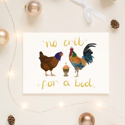 A unique christmas card lays on a table surrounded by christmas lights and ornaments. The card depicts a whimsical illustration of a chicken and cockerel dressed as Mary and Joseph and an egg cup manger containing a little egg to represent Jesus. Hand scripted yellow text read 'no crib for a bed'.