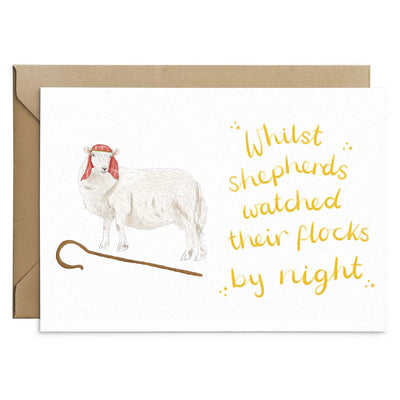 A unique sheep christmas card lays on a white background with a brown envelope behind. The card depicts a whimsical illustration of a sheep dressed as a shepherd with a shepherds crook by its feet. Hand scripted yellow text read 'Whilst shepherds watched their flocks by night'.