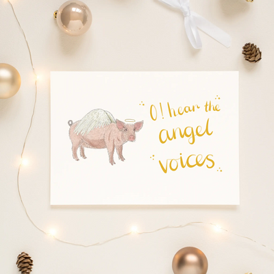 A unique christmas card lays on a table surrounded by christmas lights and ornaments. The card depicts a whimsical illustration of cute pig with angel wings and a halo.  Hand scripted yellow text reads 'O! hear the angel voices'.