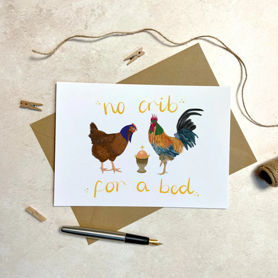 A unique christmas cards lays on a table on a brown envelope. next to a fountain pen and a ball of string. The card design depicts a whimsical illustration of nativity scene with Chicken as Mary, a cockerel as Joseph and an egg in an egg . Hand scripted yellow text reads 'no crib for a bed'.