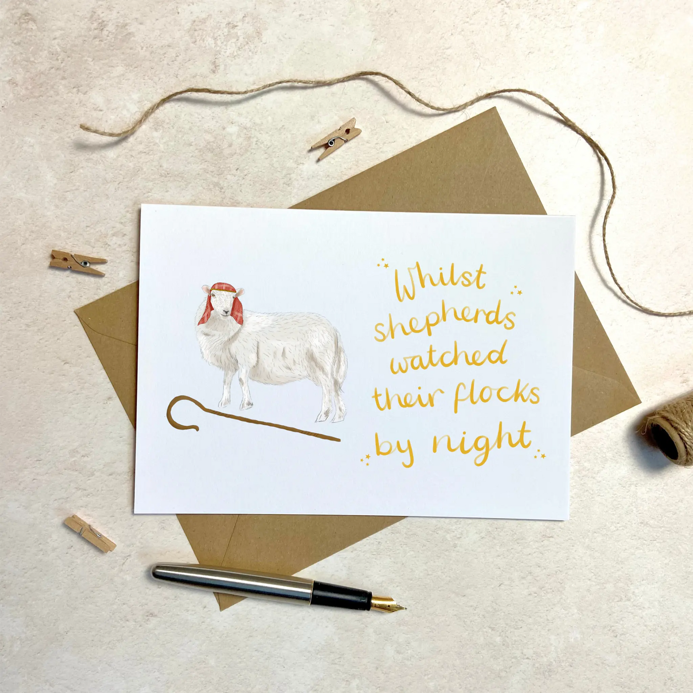 A unique christmas card lays on a table surrounded by christmas lights and ornaments. The card depicts a whimsical illustration of a sheep dressed as a shepherd with a shepherds crook by its feet. Hand scripted yellow text read 'Whilst shepherds watched their flocks by night'.