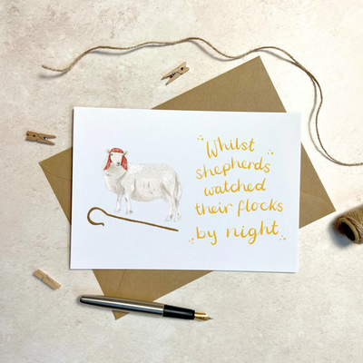 A unique christmas card lays on a table surrounded by christmas lights and ornaments. The card depicts a whimsical illustration of a sheep dressed as a shepherd with a shepherds crook by its feet. Hand scripted yellow text read 'Whilst shepherds watched their flocks by night'.