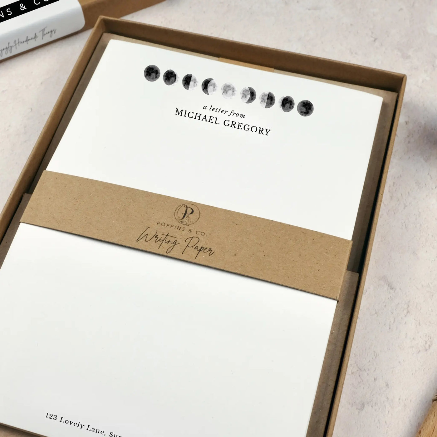 A personalised moon themed letter writing set by Poppins and co in a brown kraft luxury gift box with a cardboard band around the paper within the box. The writing paper is white and has an illustrations of the moon in different stages at the top of the page. Italic script reads 'a letter from' with the name 'Michael Gregory' typed in all caps font below it. At the bottom of the page is a typed address.