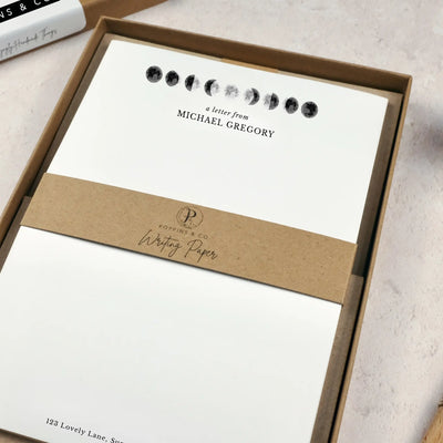 A personalised moon themed letter writing set by Poppins and co in a brown kraft luxury gift box with a cardboard band around the paper within the box. The writing paper is white and has an illustrations of the moon in different stages at the top of the page. Italic script reads 'a letter from' with the name 'Michael Gregory' typed in all caps font below it. At the bottom of the page is a typed address.