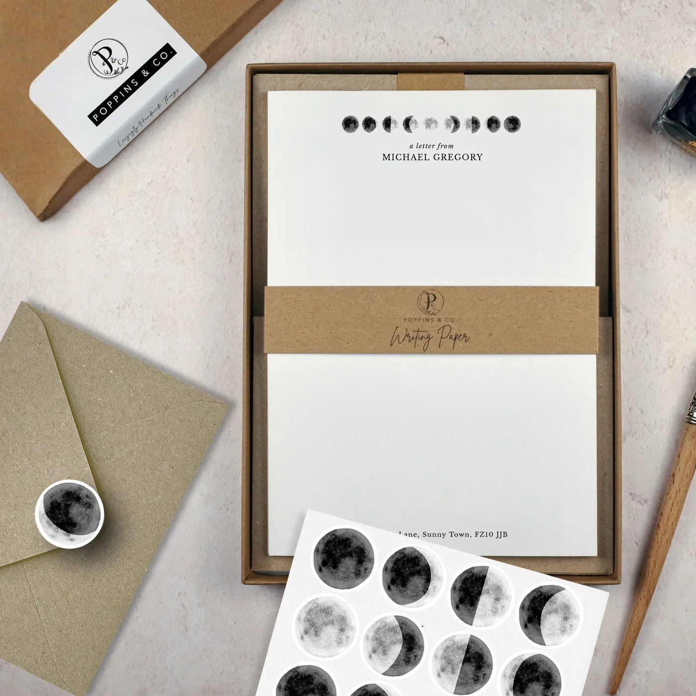 A moon themed writing paper set by in a brown kraft luxury gift box with a cardboard band around the paper within the box. The writing paper is white and has illustrations of moons in different phases at the top of the page. Italic script reads 'a letter from' with the name 'Michael Gregory' typed in all caps font below it. On top of the box lays a sheet of round envelope stickers with moon illustrations on each. To the left lays a brown kraft envelope with an envelope sticker seal on it.