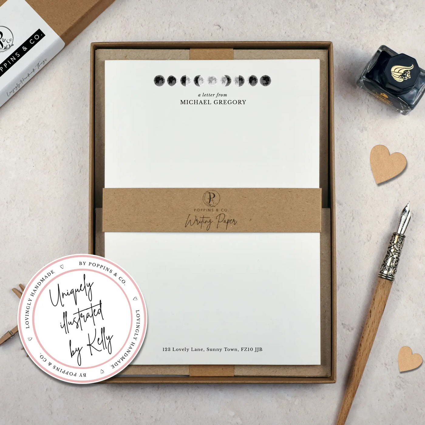 A personalised moon themed letter writing set by Poppins and co in a brown kraft luxury gift box with a cardboard band around the paper within the box. The writing paper is white and has an illustrations of the moon in different stages at the top of the page. Italic script reads 'a letter from' with the name 'Michael Gregory' typed in all caps font below it. At the bottom of the page is a typed address.