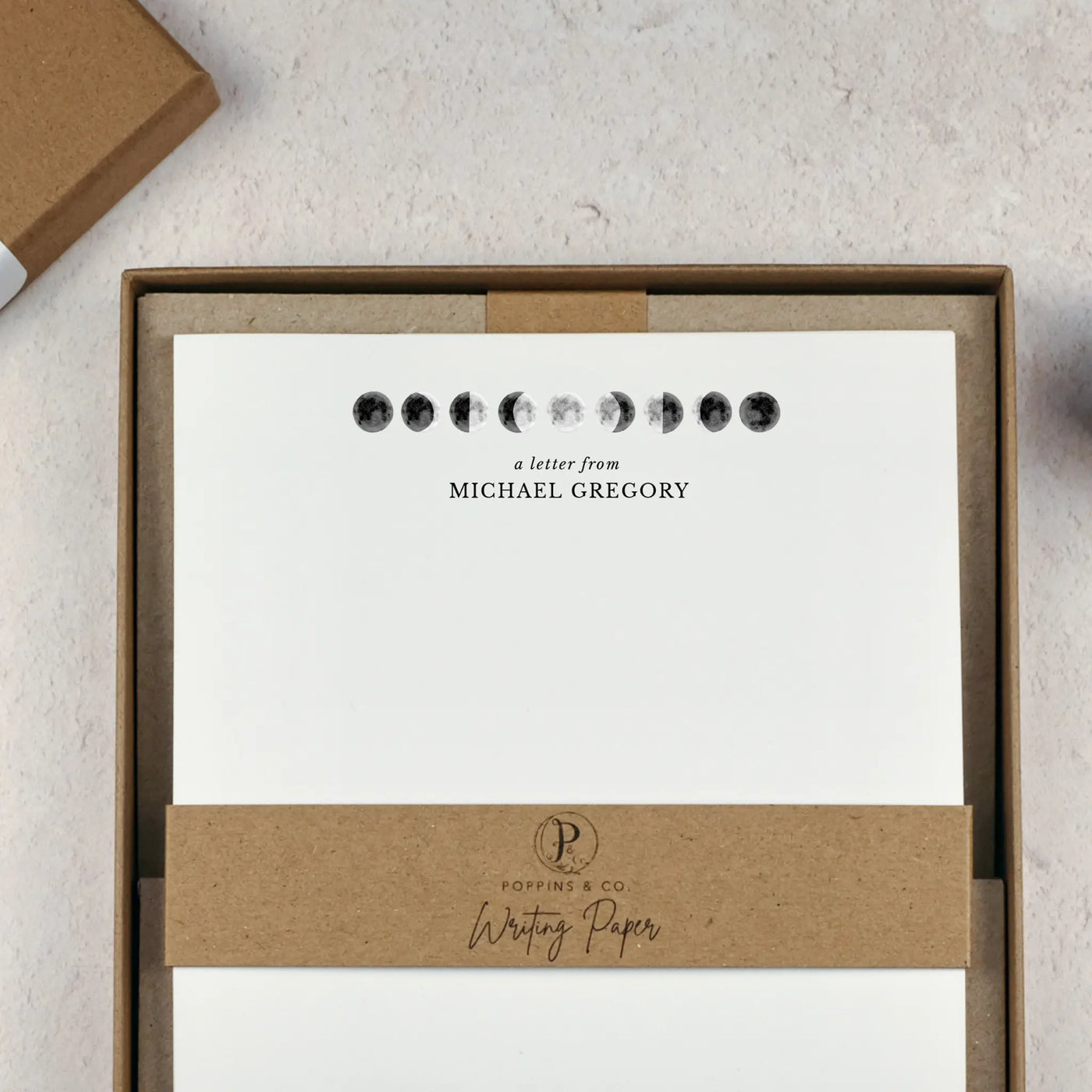 A personalised moon themed letter writing set by Poppins and co in a brown kraft luxury gift box with a cardboard band around the paper within the box. The writing paper is white and has an illustrations of the moon in different stages at the top of the page. Italic script reads 'a letter from' with the name 'Michael Gregory' typed in all caps font below it.