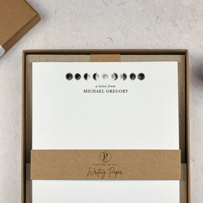 A personalised moon themed letter writing set by Poppins and co in a brown kraft luxury gift box with a cardboard band around the paper within the box. The writing paper is white and has an illustrations of the moon in different stages at the top of the page. Italic script reads 'a letter from' with the name 'Michael Gregory' typed in all caps font below it.