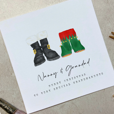 A square christmas card for grandparents featuring unique watercolour illustrations of a pair of Santa boots and elf boots side beside. Beneath the boots personalised text reads 'Nanny and Grandad - merry christmas to very special grandparents.'