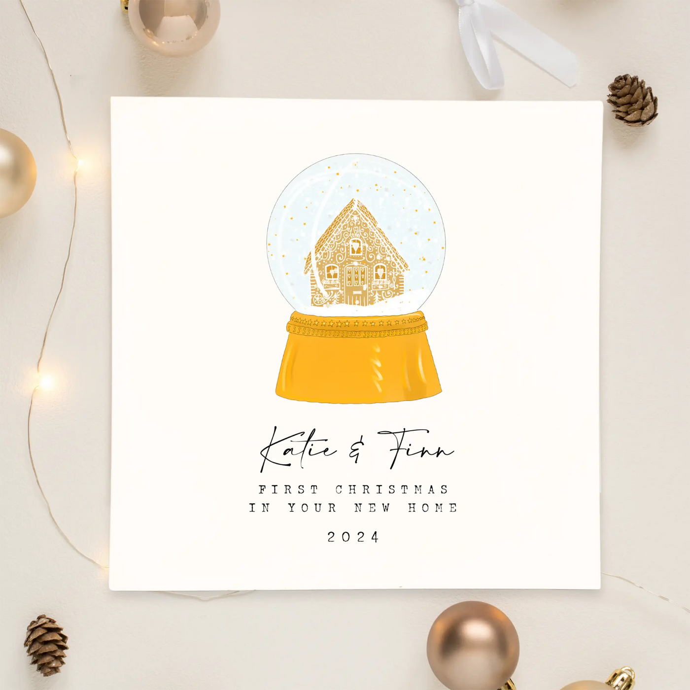 A square christmas card celebrating their first christmas in a new home. Featuring unique watercolour illustration of a snow globe containing a gingerbread house. Beneath the snow globe personalisable text reads 'Katie and Finn - first christmas in your new home 2024'