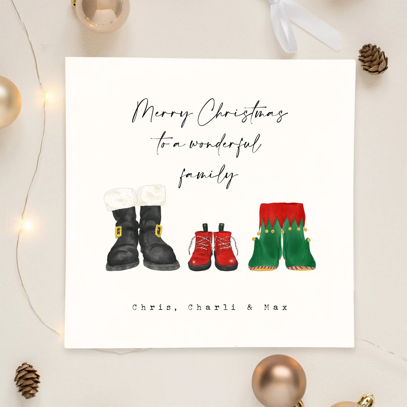 A square christmas card for a wonderful family - featuring unique watercolour illustrations of a pair of Santa boots and elf boots side beside, with baby christmas boots in-between. Personalised text reads 'Merry Christmas to a wonderful family - Chris, Charli and Max' 