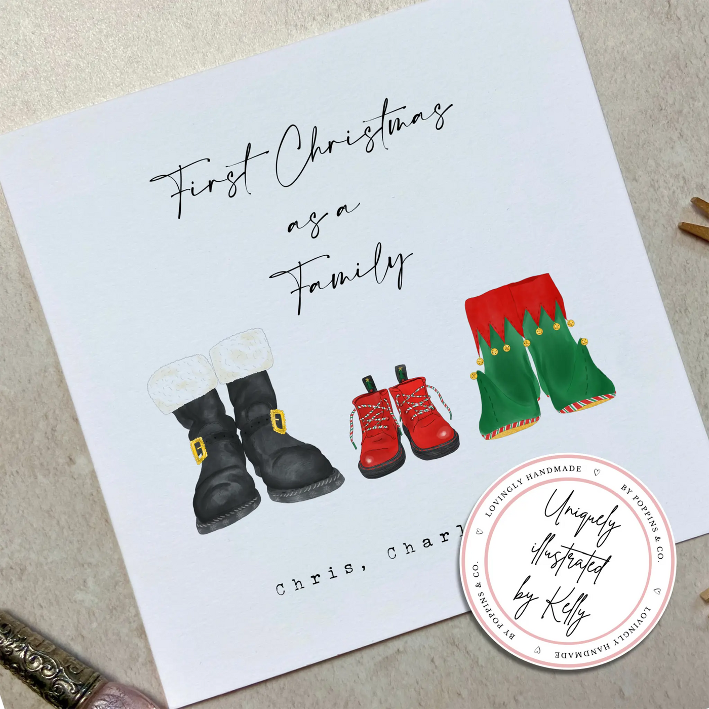 A square christmas card for their first christmas as a family - featuring unique watercolour illustrations of a pair of Santa boots and elf boots side beside, with baby christmas boots in-between. Personalised text reads 'First Christmas as a Family - Chris, Charli and Max' 