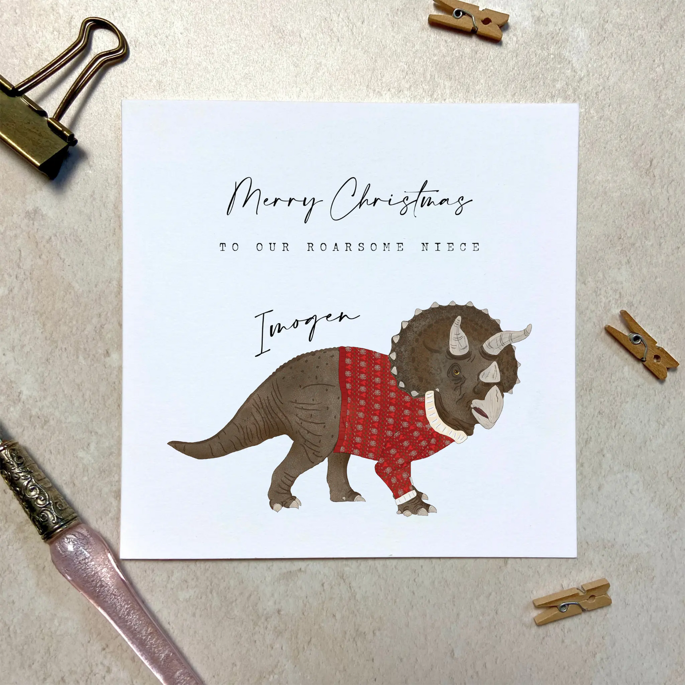 A personalised square greetings card featuring a unique illustration of triceratops in a christmas jumper and personalisable text reading 'Merry Christmas to our roar some godson Alfie'.