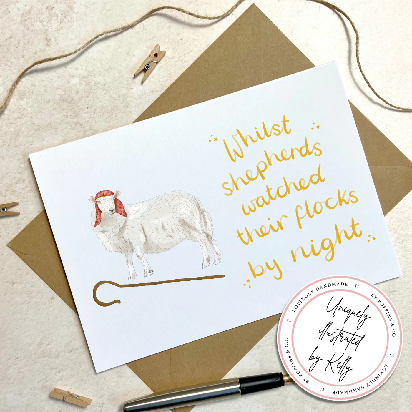 A unique christmas card lays on a table surrounded by christmas lights and ornaments. The card depicts a whimsical illustration of a sheep dressed as a shepherd with a shepherds crook by its feet. Hand scripted yellow text read 'Whilst shepherds watched their flocks by night'.