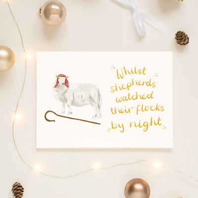 A unique christmas card lays on a table surrounded by christmas lights and ornaments. The card depicts a whimsical illustration of a sheep dressed as a shepherd with a shepherds crook by its feet. Hand scripted yellow text read 'Whilst shepherds watched their flocks by night'.