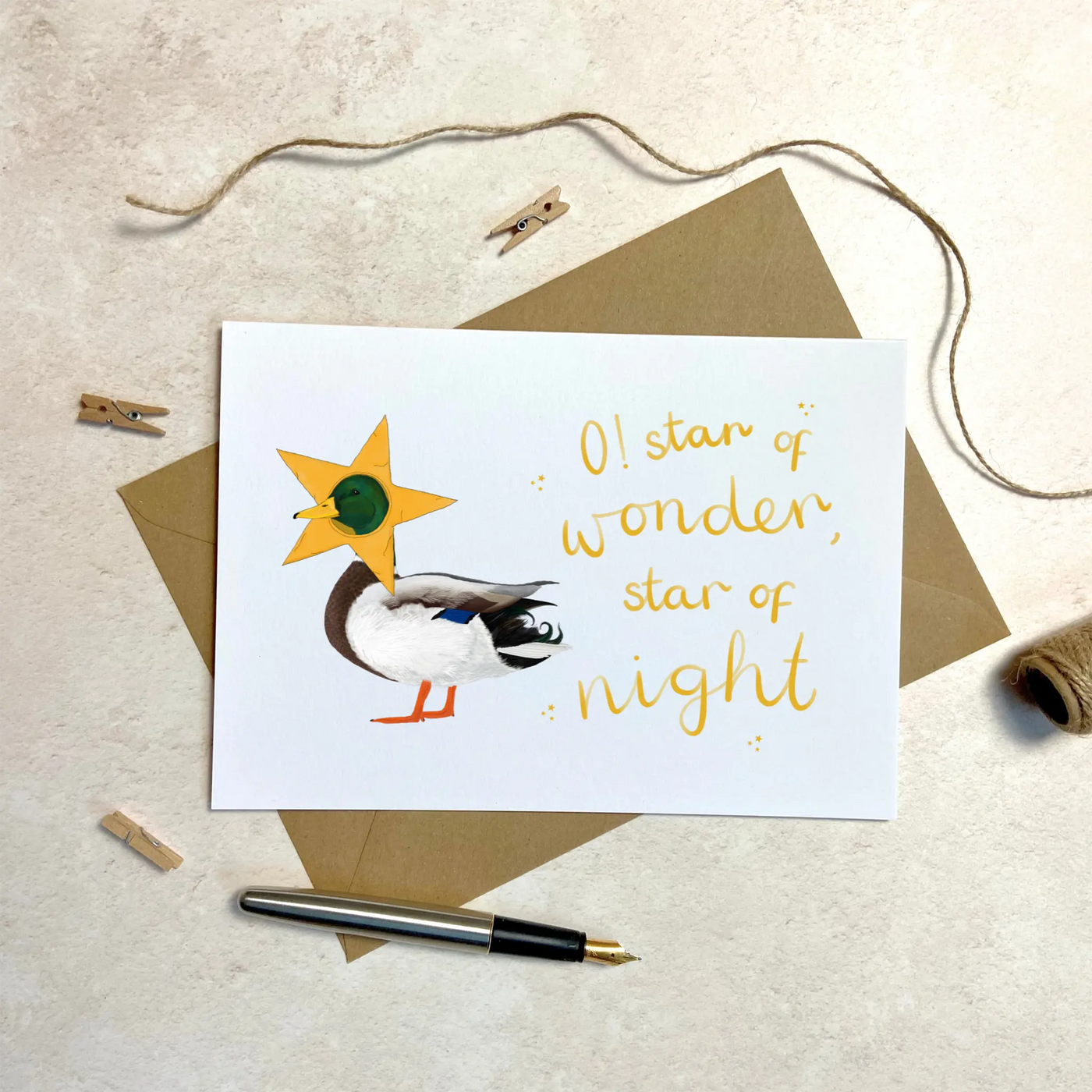 A unique christmas card lays on a table surrounded by christmas lights and ornaments. The card depicts a whimsical illustration of cute duck with a star costume around its head.  Hand scripted yellow text reads 'O! star of wonder, star of night'.