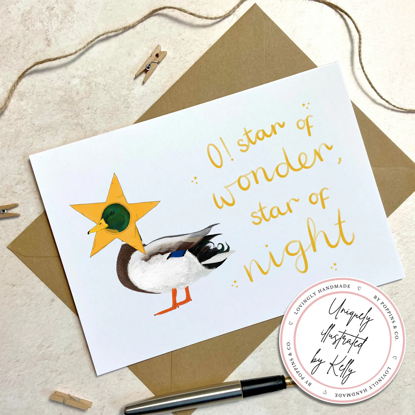 A unique christmas card lays on a table surrounded by christmas lights and ornaments. The card depicts a whimsical illustration of cute duck with a star costume around its head.  Hand scripted yellow text reads 'O! star of wonder, star of night'.