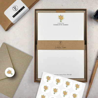 A letter writing set by in a brown kraft luxury gift box with a cardboard band around the paper within the box. The writing paper is white and has a small bunch of yellow sunflowers illustrated at the top of the page. Italic script reads 'a letter from' with the name 'Charlotte Harris' typed in all caps font below it. On top of the box lays a sheet of round envelope stickers with the same sunflower illustration on each. To the left lays a brown kraft envelope with an envelope sticker seal on it.