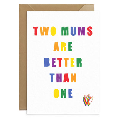 Two mums are better than one greetings card. A white greetings card with rainbow writing on in red orange yellow green blue and violet. Two small rainbow hearts on bottom right corner or card. Behind the card you can see the corners of a brown craft envelope. Greetings Card by Poppins and Co.